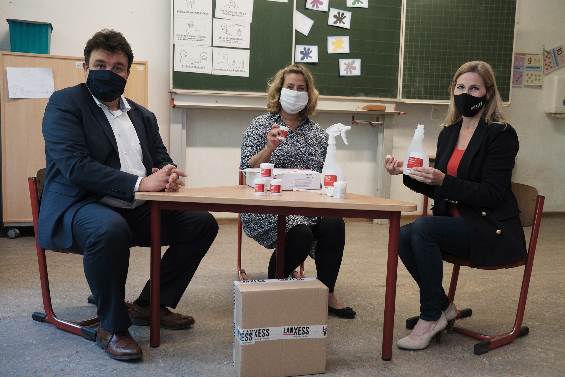 On behalf of all Krefeld schools, Nina Hasenkamp, head of the LANXESS education initiative, hands over a package containing the disinfectant Rely+On Virkon to the headmistress of GGS im Krähenfeld, D. Schrader, and the deputy of the city of Krefeld, Markus Schön.