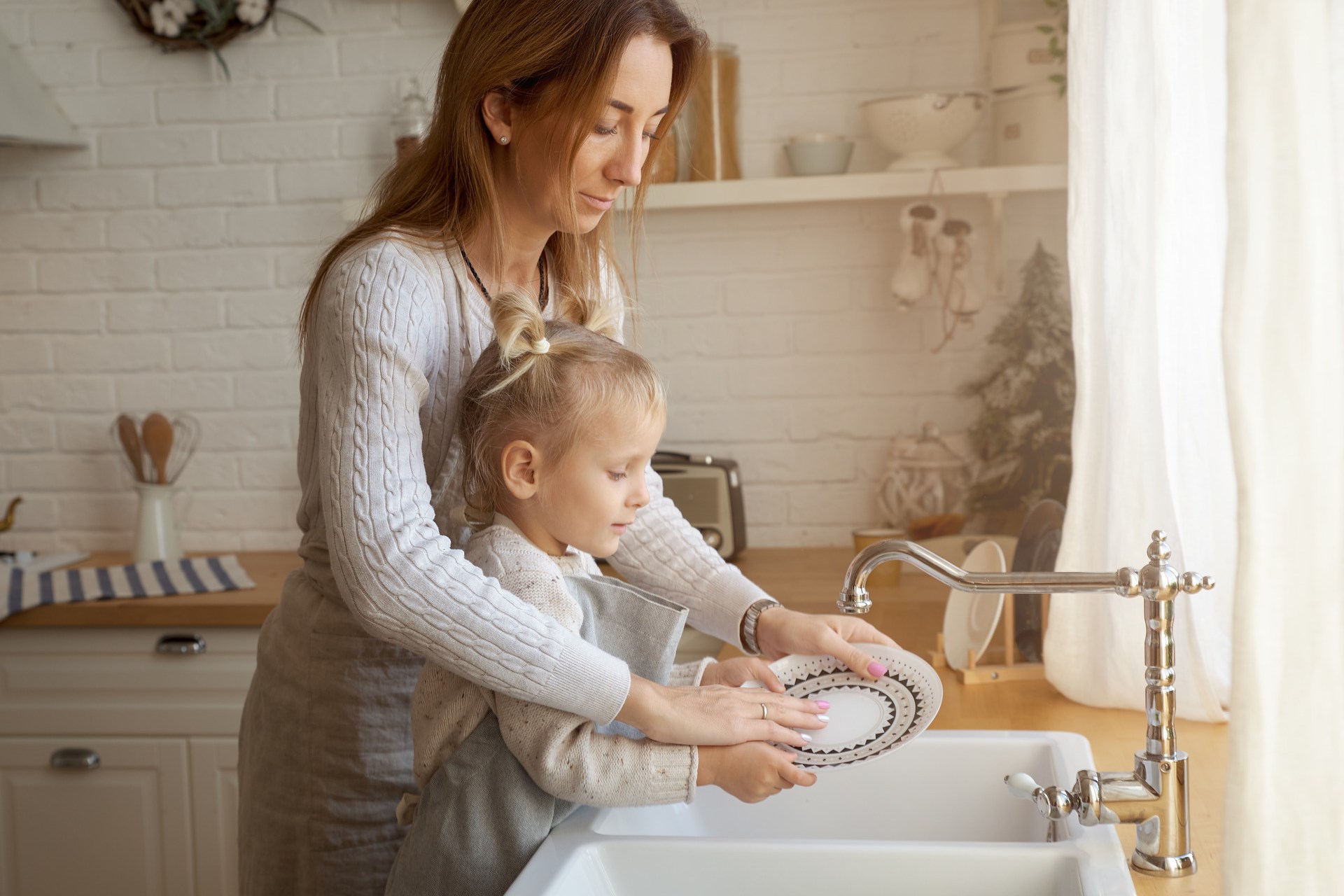 A woman does the dishes with a child in the kitchen.