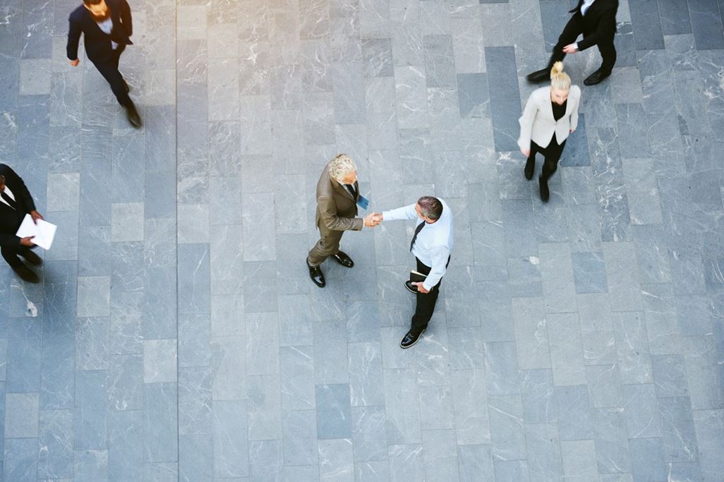 High angle view of two businessmen shaking hands together while colleagues walk around them in the lobby of a busy modern office building