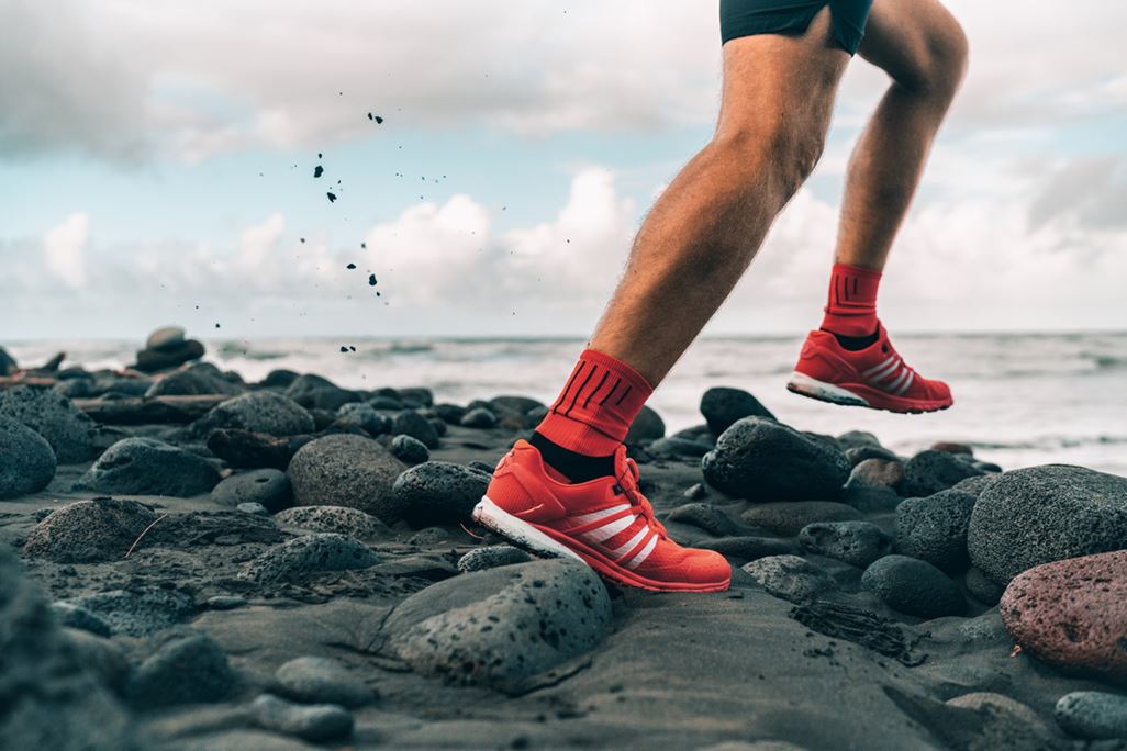 Running shoes of athlete man jogging on beach. Difficult terrain with wet rocks for training outdoors. Runner exercising.