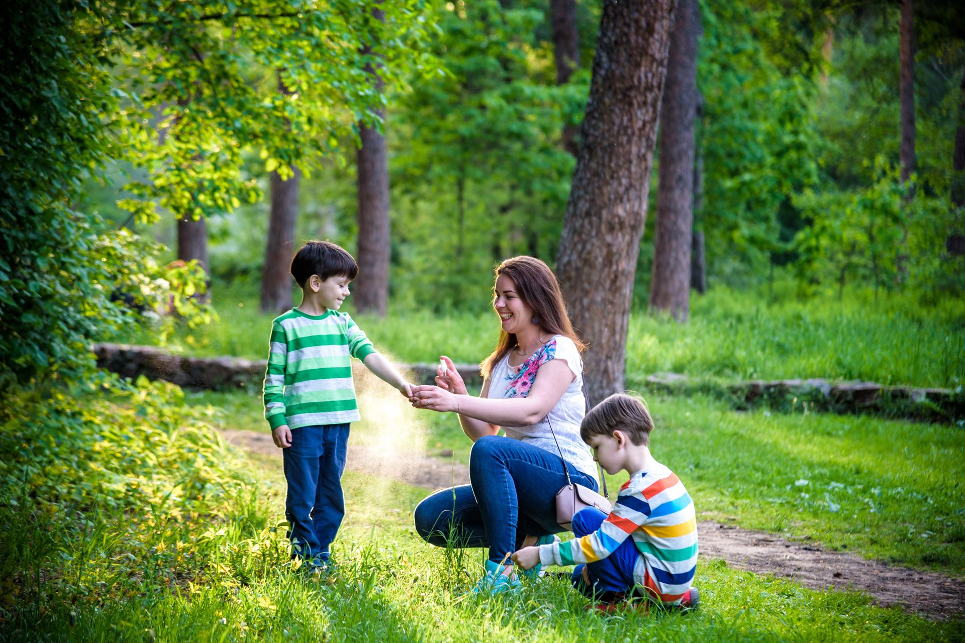 Young woman mother applying insect repellent to her two son before forest hike beautiful summer day or evening. Protecting children from biting insects at summer. Active leisure with kids,