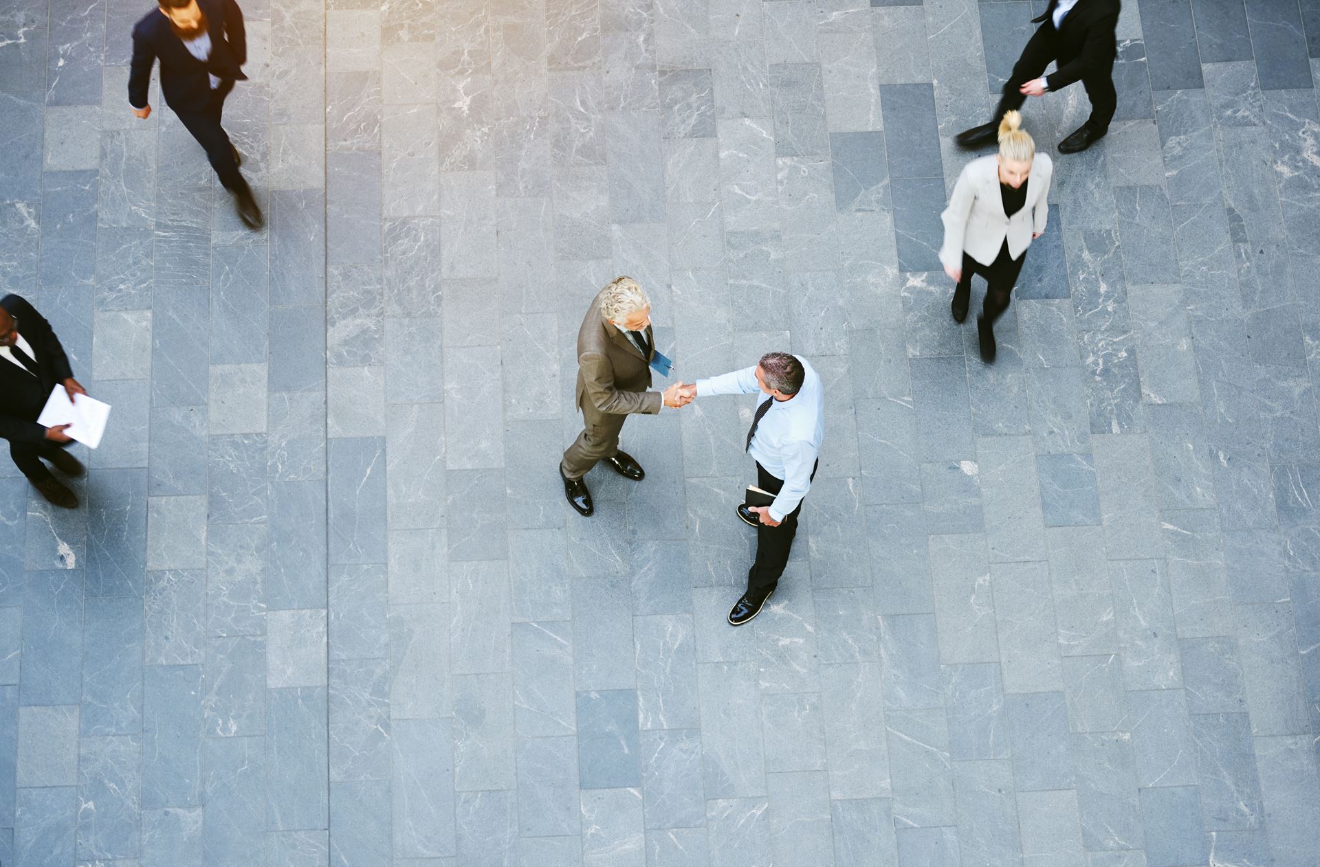 High angle view of two businessmen shaking hands together while colleagues walk around them in the lobby of a busy modern office building
