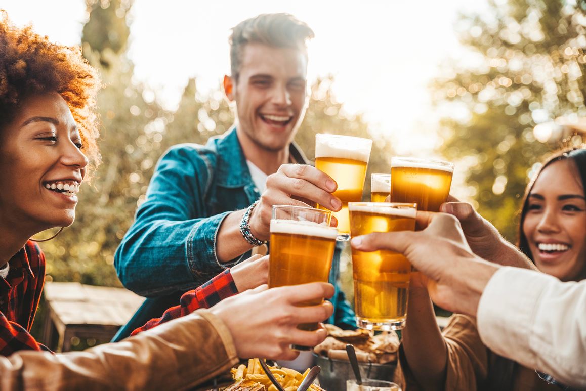 Group of multi ethnic friends having backyard dinner party together - Diverse young people sitting at bar table toasting beer glasses in brewery pub garden - Happy hour, lunch break and youth concept