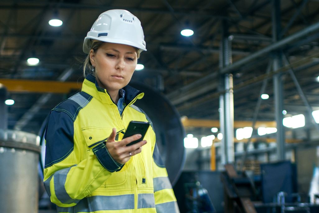 Female Industrial Worker in the Hard Hat Uses Mobile Phone While Walking Through Heavy Industry Manufacturing Factory. In the Background Various Metalwork Project Parts Lying