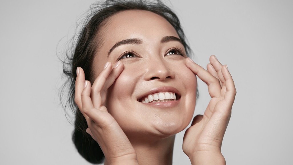 Skin care. Woman with beauty face touching healthy facial skin portrait. Beautiful smiling asian girl model with natural makeup touching glowing hydrated skin on blue background closeup